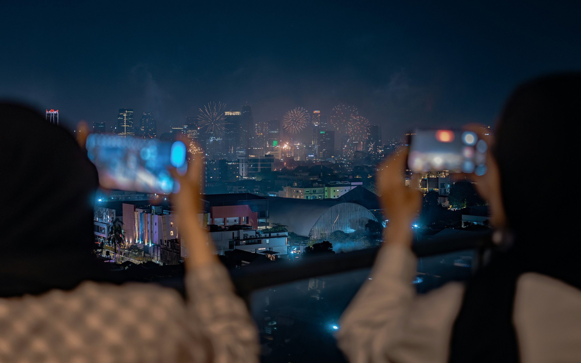 Two people taking pictures of fireworks over a cityscape